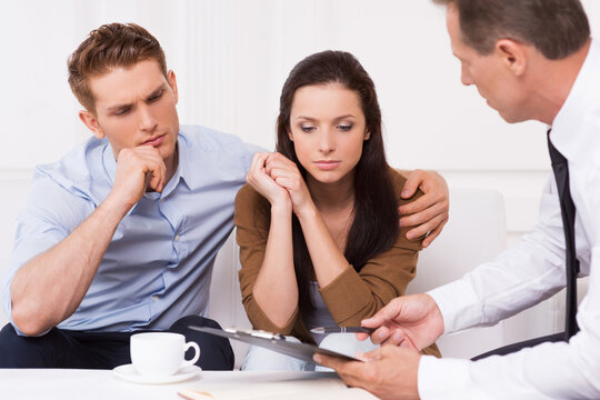 Expert advice. Thoughtful young couple sitting on the couch while confident financial advisor explaining something and pointing clipboard