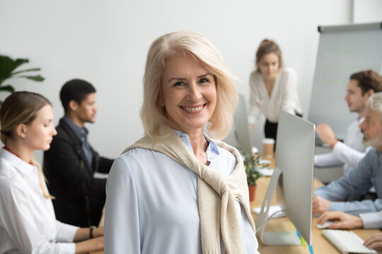 Smiling female aged company executive or team leader looking at camera, happy senior businesswoman teacher coach posing with office people at background, friendly older woman boss head shot portrait